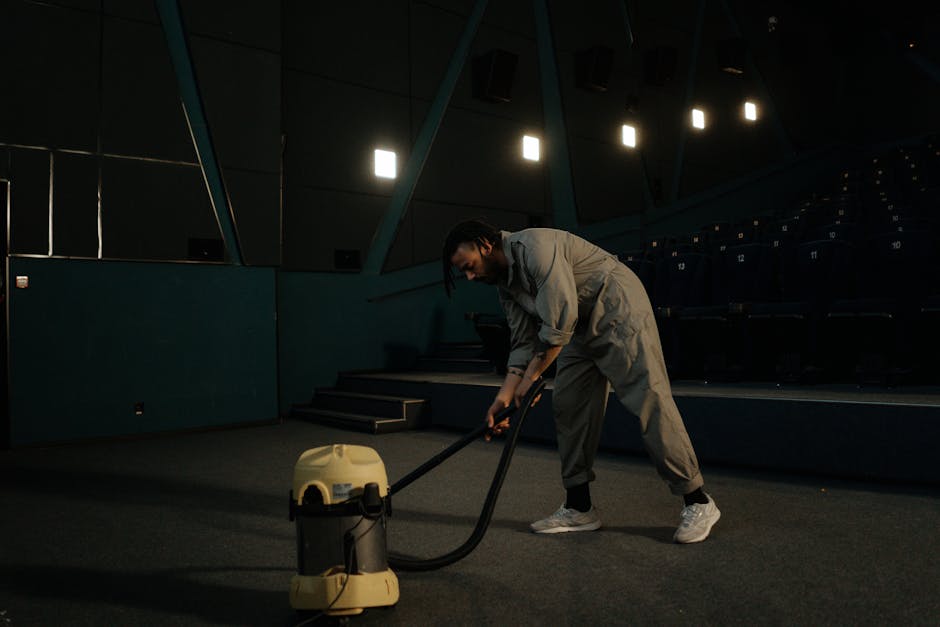 A person wearing grey overalls and white sneakers is operating a yellow commercial vacuum cleaner on a dark, carpeted auditorium floor with tiered seating. The room is dimly lit by overhead lights, an