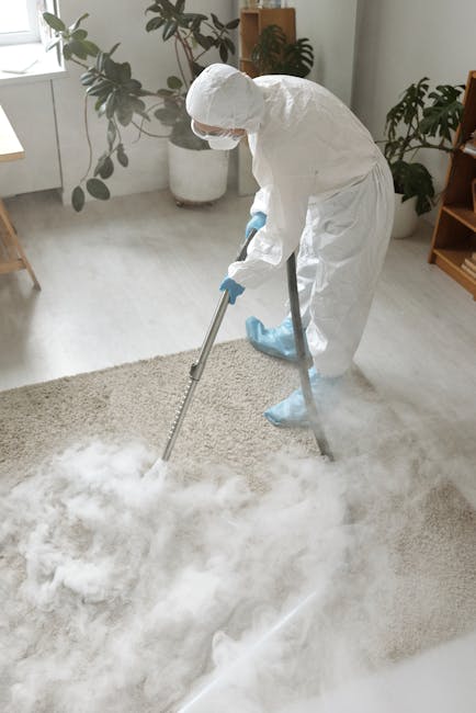 A person wearing grey overalls and white sneakers is operating a yellow commercial vacuum cleaner on a dark, carpeted auditorium floor with tiered seating. The room is dimly lit by overhead lights, an