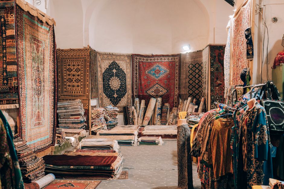 At an outdoor market, several intricately patterned Persian-style rugs featuring elaborate gold, black, green, pink, and beige designs are displayed on tables. The rugs are clean and well-maintained, 